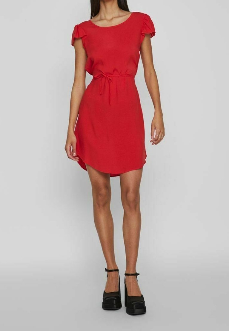 Vestido rojo con mangas cortas con volantes, cinturilla fruncida y un dobladillo ligeramente asimétrico. Lleva unos zapatos de plataforma negros.