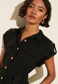 Zwarte button-up jurk met korte opgerolde mouwen, twee borstzakken en gouden bloemenknopen. De stof lijkt glad en lichtgewicht.