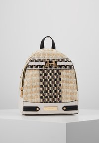 Sac à dos en tissu beige avec accents noirs, présentant un motif texturé, une quincaillerie dorée et une plaque de logo minimaliste. Compartiments zippés et poignée de transport.