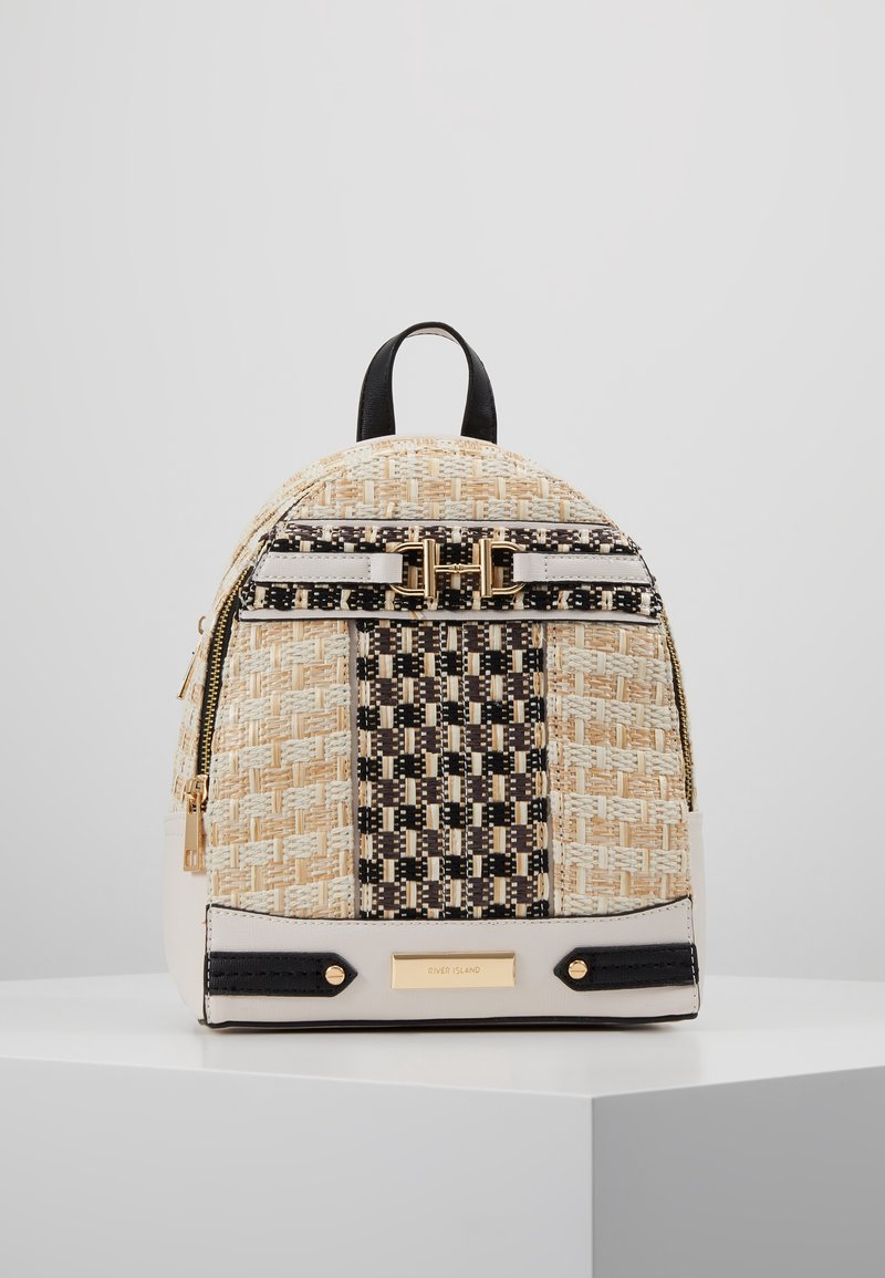Sac à dos en tissu beige avec accents noirs, présentant un motif texturé, une quincaillerie dorée et une plaque de logo minimaliste. Compartiments zippés et poignée de transport.