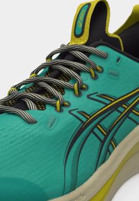 Teal Asics løpesko med svarte og gule detaljer, stripete svarte og hvite skolisser, og polstret såle synlig mot hvit bakgrunn.
