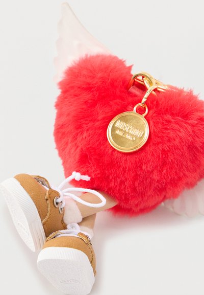 Porte-clés en forme de cœur rouge duveteux avec une étiquette en or et des jambes de poupée attachées, portant des chaussures marron et des chaussettes blanches, sur fond blanc.