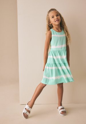 Junges Mädchen mit langen lockigen Haaren, das ein ärmelloses, mintgrünes und weiß gestreiftes Kleid sowie weiße Sandalen mit Schnallen trägt und auf einem Bein steht.