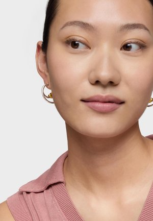 Giovane con pelle liscia, indossa una canottiera rosa e orecchini a cerchio d'argento con dettagli dorati, guarda di lato.