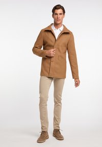 Jachetă din lână maro, cu guler cu margini, buzunare frontale și închidere cu nasturi. Pantaloni slim-fit bej și pantofi din suede bej completează ținuta.