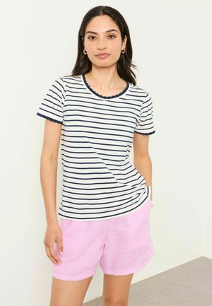 Young woman with dark hair wearing a white short-sleeve navy-striped shirt and light pink shorts, standing against a white wall.