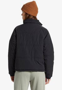 Sort puffer-jakke med høj krave, quiltet tekstur og løs pasform. Bæres med en brun strikket hue. Synlige søm-detaljer.