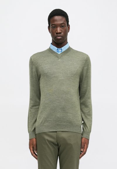 Uomo che indossa un maglione verde oliva a V sopra una camicia colletto blu chiaro e pantaloni verdi oliva, in piedi contro uno sfondo bianco uniforme.