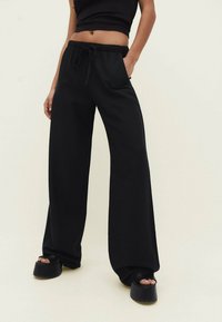 Pantalon noir large en tissu doux avec une ceinture élastique et un cordon de serrage, doté de poches latérales et d'une texture lisse.