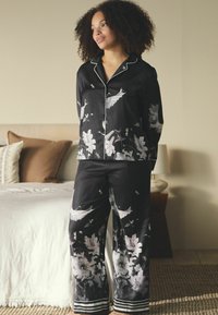 Fekete szatén pizsamaszett virág- és madármintákkal, fehér szegéllyel a galléron és a mandzsettán. Bő fazonú nadrág csíkos szegéllyel.