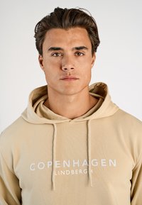 Felpa beige con tasca a marsupio frontale, cappuccio con coulisse e scritta bianca "COPENHAGEN LINDBERGH". Tessuto morbido e strutturato.