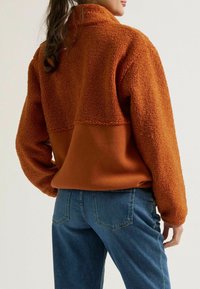 Brauner Fleece-Pullover mit hohem Kragen, der im oberen Teil mit strukturiertem Stoff und im unteren Bereich mit glattem Material ausgestattet ist. Kombiniert mit blauen Jeans.