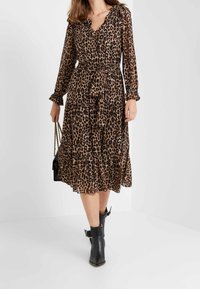 Femme portant une robe léopard jusqu'aux genoux avec une ceinture à la taille, tenant un sac à main noir et portant des bottes noires à la cheville avec des boucles.