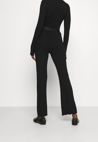 Czarny prążkowany kombinezon z długimi rękawami, dopasowaną sylwetką i rozszerzanymi spodniami. Posiada wiązany w talii pasek oraz postrzępione detale u dołu. Nosi się z czarnymi butami.