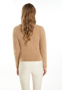Teddybeige fluffy trui met een ronde halslijn en lange mouwen, gecombineerd met glanzende witte broeken. De achterkant toont een zachte textuur en een getailleerd ontwerp.