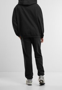 Person trägt schwarzen Kapuzenpullover und passende Jogginghose, läuft auf weißem Boden weg, graue und weiße Sneakers sichtbar.