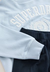 Šviesiai mėlynas džemperis su baltais siuvinėtais užrašais ir tamsiai mėlynos kelnės, turinčios ribotuotus rankogalius ir elastingą juosmenį.