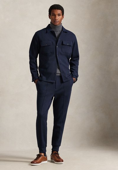 Μπλε πουκάμισο-σακάκι από μάλλινο μείγμα με δύο τσέπες στο στήθος, συνδυασμένο με τσάκιζα παντελόνια στο ίδιο ύφος και καφέ δερμάτινα αθλητικά παπούτσια. Υφασμένο ζιβάγκο με ανάγλυφη υφή από μέσα.