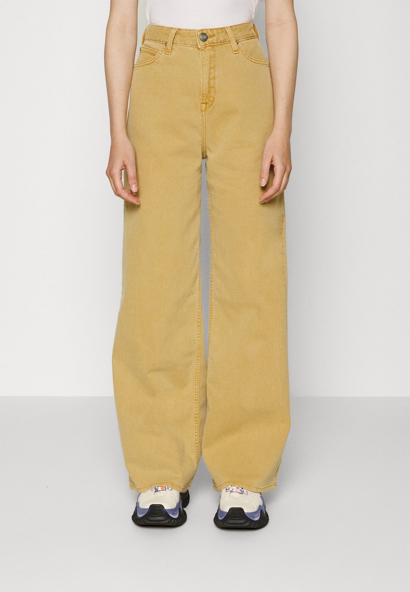 Personne portant un jean jaune moutarde à jambes larges avec des baskets blanches et noires, debout devant un fond clair uni.