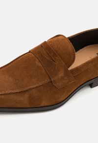 Loafer din suede maron, cu toc plat, având un accent cusut pe vârful pantofului și un finisaj texturat, potrivit pentru ținute casual.
