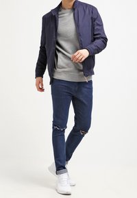 Námořnická bombardovací bunda přes šedou mikinu, doplněná o potrhané tmavé džíny a bílé tenisky. Neformální styl s vrstveným vzhledem.