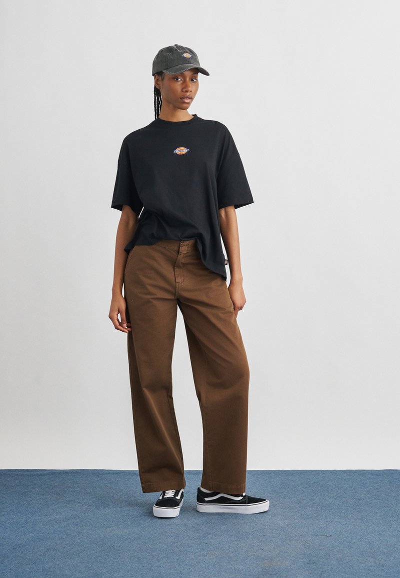 Giovane donna che indossa una T-shirt nera con logo, pantaloni marroni a gamba larga, sneakers nere e bianche, e un cappellino grigio, in piedi su un tappeto blu.