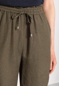 Olijfgroene broek met elastische taille en een strik in de veter, gedragen met een donker shirt, waarbij een deel van de arm en hand van een persoon zichtbaar is.