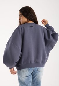 HallGrey oversized-sweatshirt, mille on turske varrude, ribitud krae ja äär, taga on väike logo. Kombineeritud helesinise denimistega.