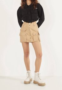 Černá krátká bunda přes béžovou cargo sukni se zipem vpředu a více kapsami. Modelka má na sobě bílé šněrovací boty s průhlednou podrážkou.