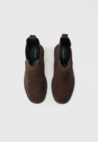 Ruskeat mokkanahkaiset nilkkurit, joissa on joustavat sivupaneelit, mustat kumipohjat ja vetoremmit helpottamaan pukemista. Tasapohjaiset, pyöreällä kärjellä varustetut kengät.