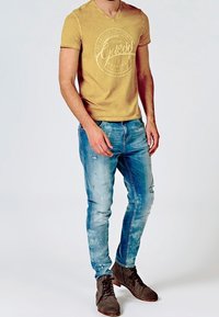 Mand iført en gul grafisk T-shirt, falmede blå slidte jeans og brune ruskinds snørebåndsstøvler står på en ensfarvet baggrund.