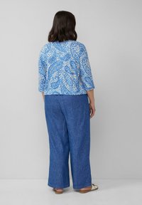 Blau-weiße gemusterte Bluse mit Dreiviertelärmeln, kombiniert mit lockeren, strukturierten blauen Hosen. Flache Sandalen runden das Outfit ab.