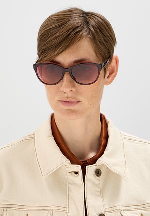 Persona con capelli corti castani che indossa occhiali da sole con lenti marroni e una giacca beige sopra una camicia marrone su uno sfondo semplice.