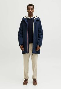 Marineblaue wasserdichte Jacke mit gefütterter Kapuze, Reißverschluss und Akzentfarben. Kombiniert mit beigen Hosen und braunen Schuhen.