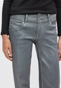 Hallid tikandiga teksapüksid kitsas lõikega, millel on kaks eest taskut, metalist nööbid ja sile tekstuur. Stiilitud musta pikkade varrukatega topiga.