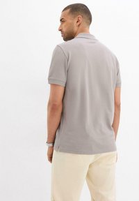 Polo grigio a maniche corte, con texture liscia e spacchi laterali. Indossato con pantaloni beige chiaro, che mostrano un design semplice e casual.
