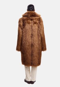 Brun kunstpelsfrakke med høj krave, lange ærmer, løs pasform og struktureret overflade. Båret over cremefarvede bukser.