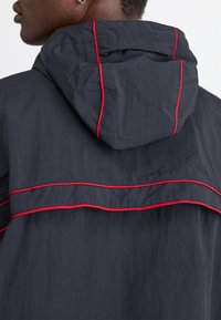 Chaqueta con capucha negra que presenta acentos de costura roja y un diseño en capas en la parte posterior. Tejido texturizado y ligero.