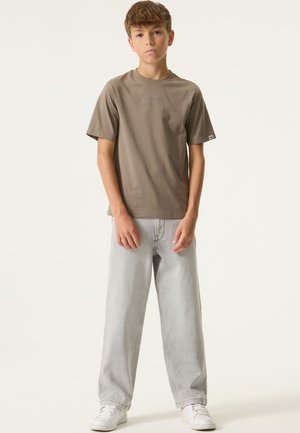 Bruine katoenen T-shirt met korte mouwen en de tekst "FUTURE" op de voorkant, gecombineerd met lichtgrijze wijde spijkerbroek en witte sneakers.