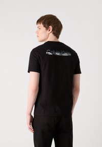 Melnas kokvilnas t-krekls ar daudzkrāsu "Calvin Klein" logotipu, kas drukāts mugurā. Īsas piedurknes un klasiskā apaļā kakla izgriezuma dizains.