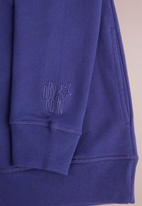 Felpa viola con polsini e orlo a coste. Presenta un logo "DRY KORN" in rilievo e dettagli sulla tasca. Tessuto morbido e testurizzato.