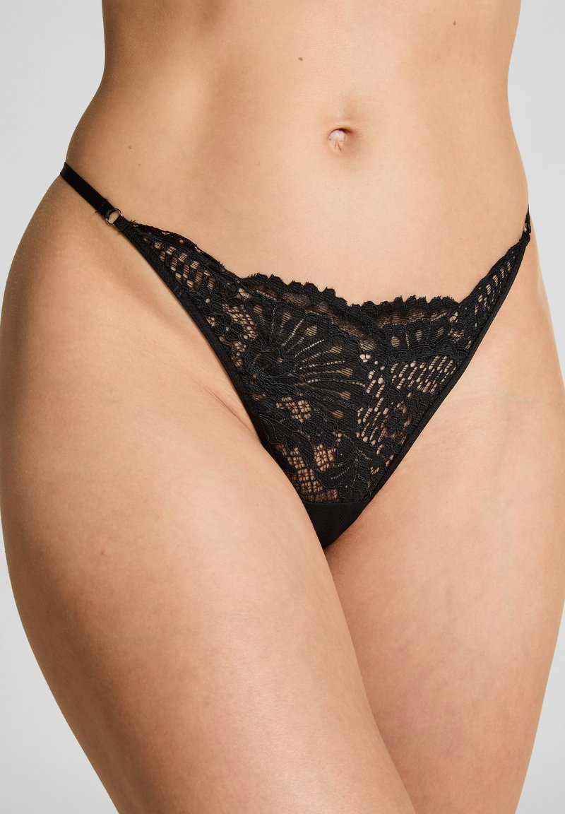 Schwarzer Spitze-Thong mit einem komplizierten Blumenmuster, ausgestattet mit dünnen Riemen und einem nahtlosen Design. Das Material wirkt weich und zart.