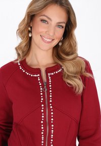 Sudadera roja con cremallera, hecha de mezcla de algodón, con adornos de perlas a lo largo de la cremallera y el escote, con una textura suave y un diseño entallado.