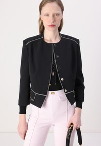 Černá zkrácená bunda s bílým lemem, kulatým výstřihem a zapínáním na knoflíky. Model má na sobě růžové tailored kalhoty. Obsahuje zlaté detaily a kapsy.