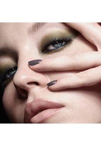 Gros plan sur un visage avec un fard à paupières vert fumé, une peau claire et des lèvres nudes. Les ongles sont courts, brillants et de couleur gris foncé.