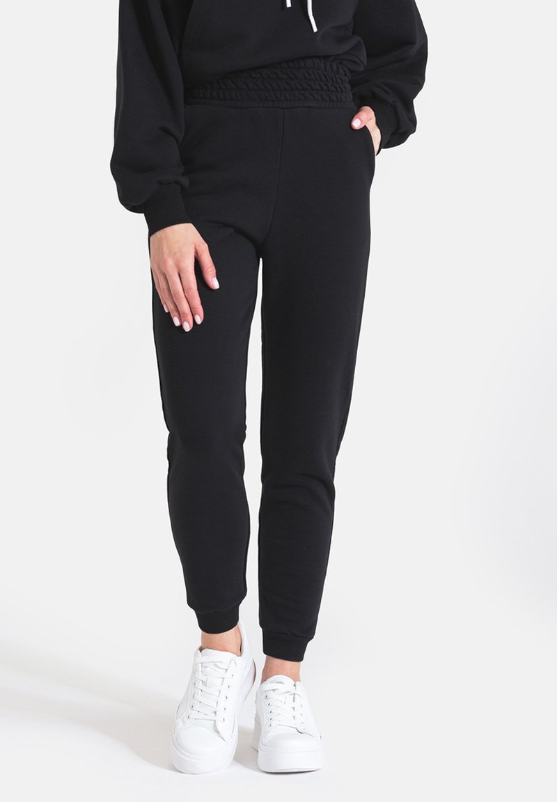 Czarne dresowe spodnie z marszczonym pasem, zwężanym krojem nogawki i kieszeniami, w połączeniu z białymi sneakersami. Miękka, elastyczna tekstura materiału.