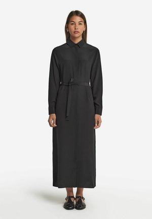 Robe noire à manches longues en tissu lisse avec un col, une taille cintrée et une silhouette droite. Portée avec des sandales noires.