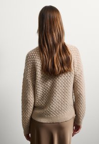 Maglione lavorato a maglia beige con un motivo testurizzato, orlo e polsini a coste. Maniche lunghe, vestibilità comoda, indossato sopra una gonna marrone liscia.