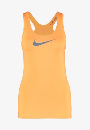 Donna che indossa una canotta Nike arancione, pantaloncini da ciclismo neri e scarpe da corsa nero-verdi che cammina davanti a uno sfondo semplice.