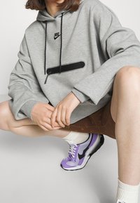 Sudadera gris con detalles en negro y un bolsillo con cremallera. Combinada con pantalones cortos marrones con un patrón y zapatillas Nike en púrpura y blanco.
